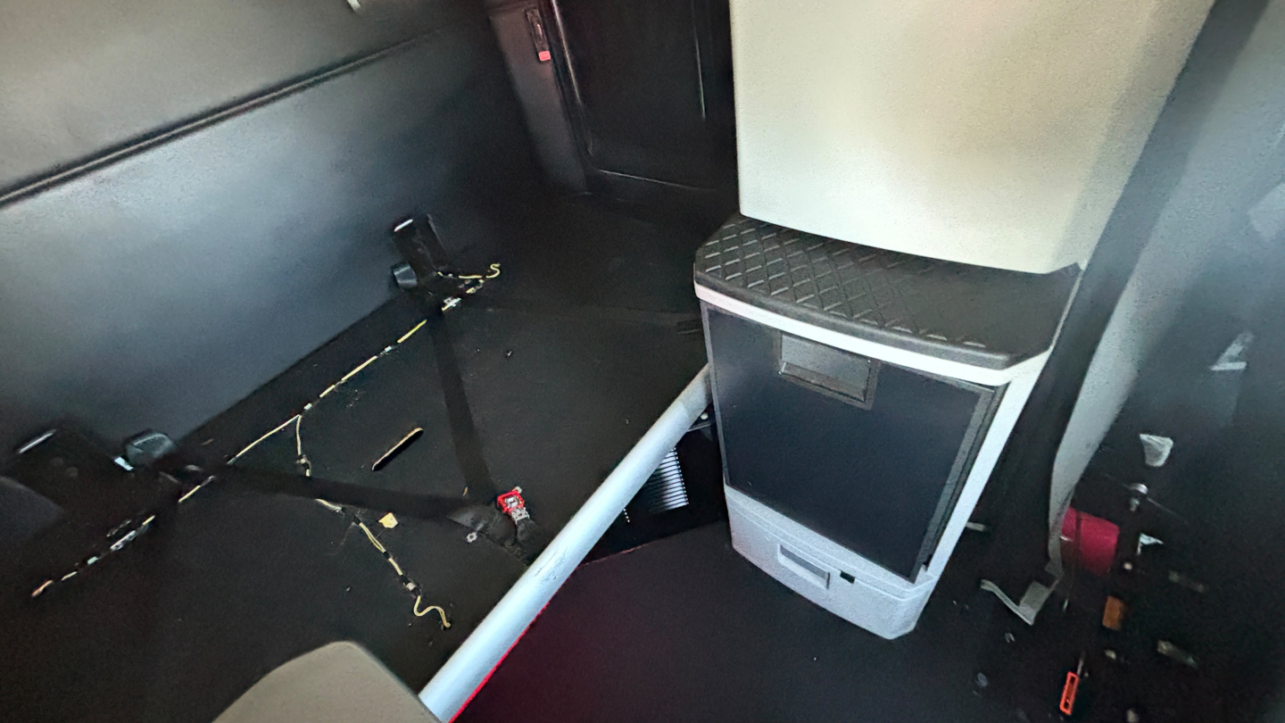 Cab interior of a Kenworth truck showing a space with scattered tools, floor debris, and a small auxiliary cabinet or cooler mounted near the aisle.