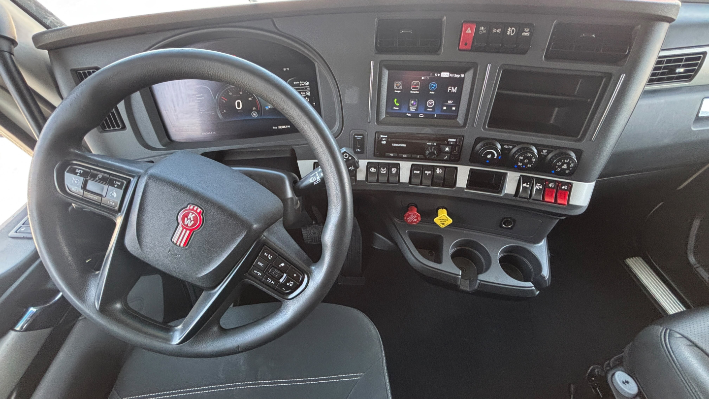 Truck interior dashboard of a Kenworth T680 showing steering wheel with logo, center console with touchscreen display, various gauges, climate controls, and gear shifter.