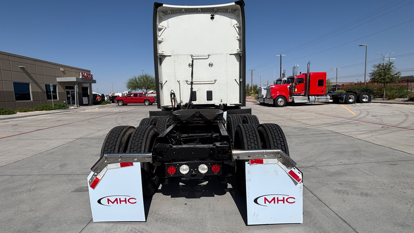 Truck rear view showing fifth wheel, dual rear axles, and white MHC mudflaps with red MHC logos; cab in background; clear blue sky.