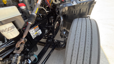Kenworth truck engine bay and front suspension view in-cabin area showing hoses, wiring, and label stickers.