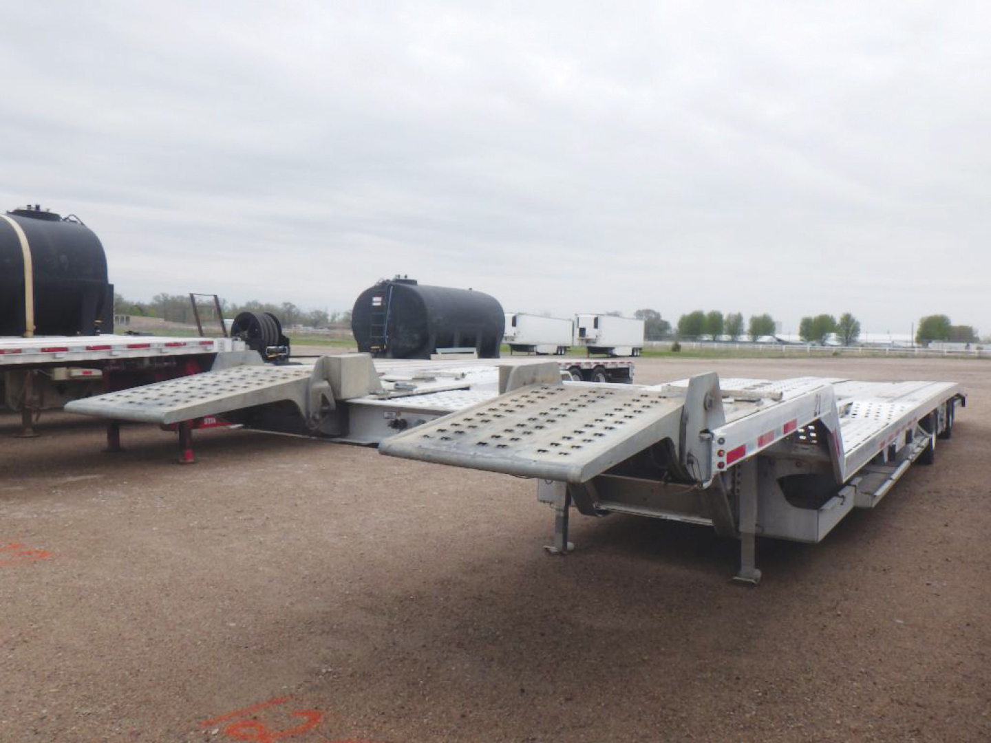 Aluminum drop-deck trailer with multiple loading ramps and a flatbed section, parked on a gravel lot with other trailers and equipment in the background.