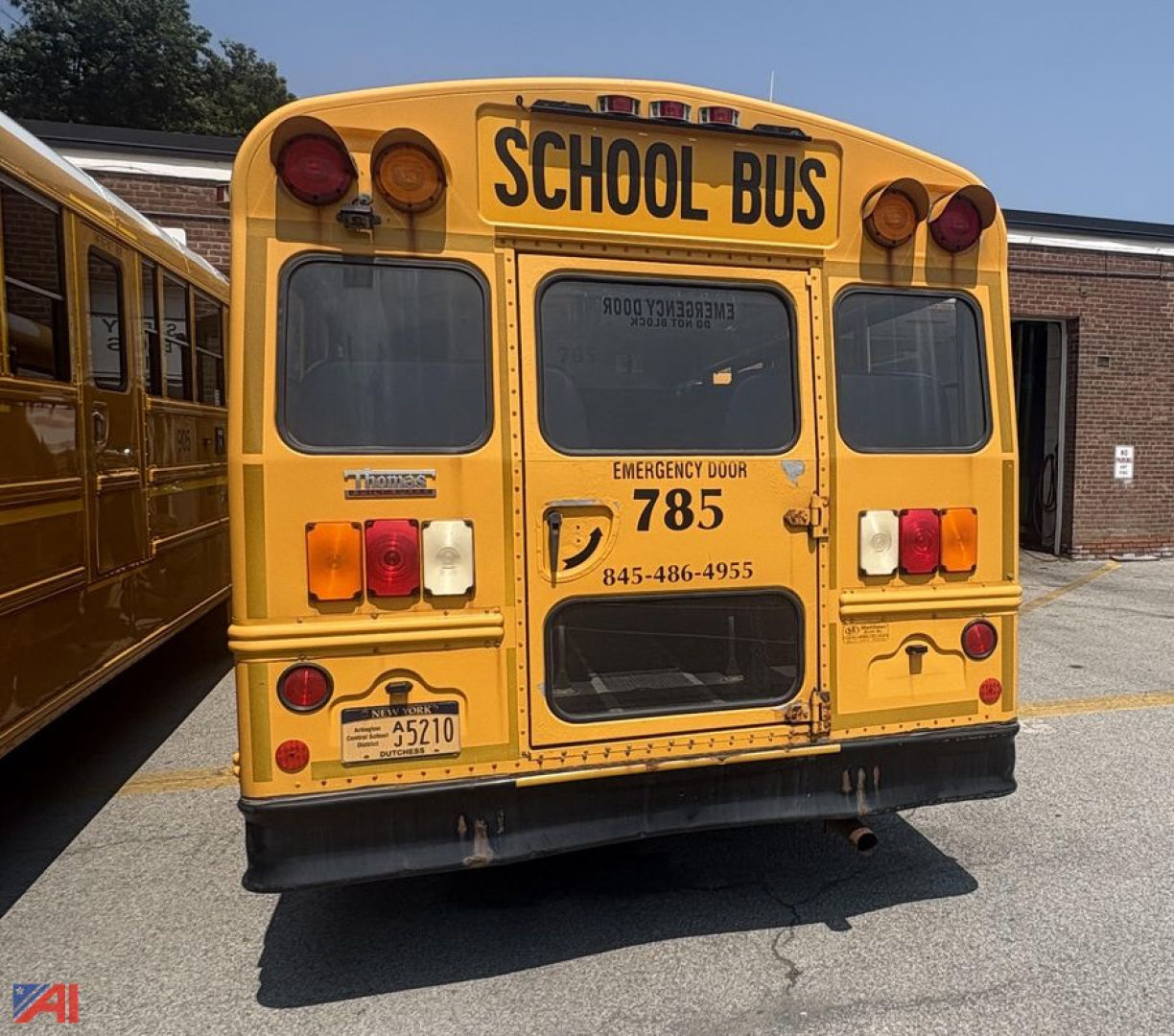 Rear view of a yellow school bus with 'SCHOOL BUS' signage, red/amber lights, rear door, and emergency exit with phone number visible.