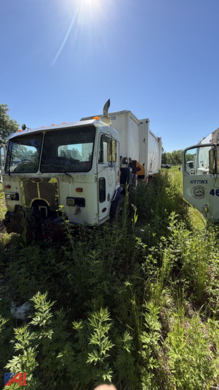 Truck cab and side-arm garbage body in overgrown lot, Peterbilt 320 side-arm refuse truck visible.