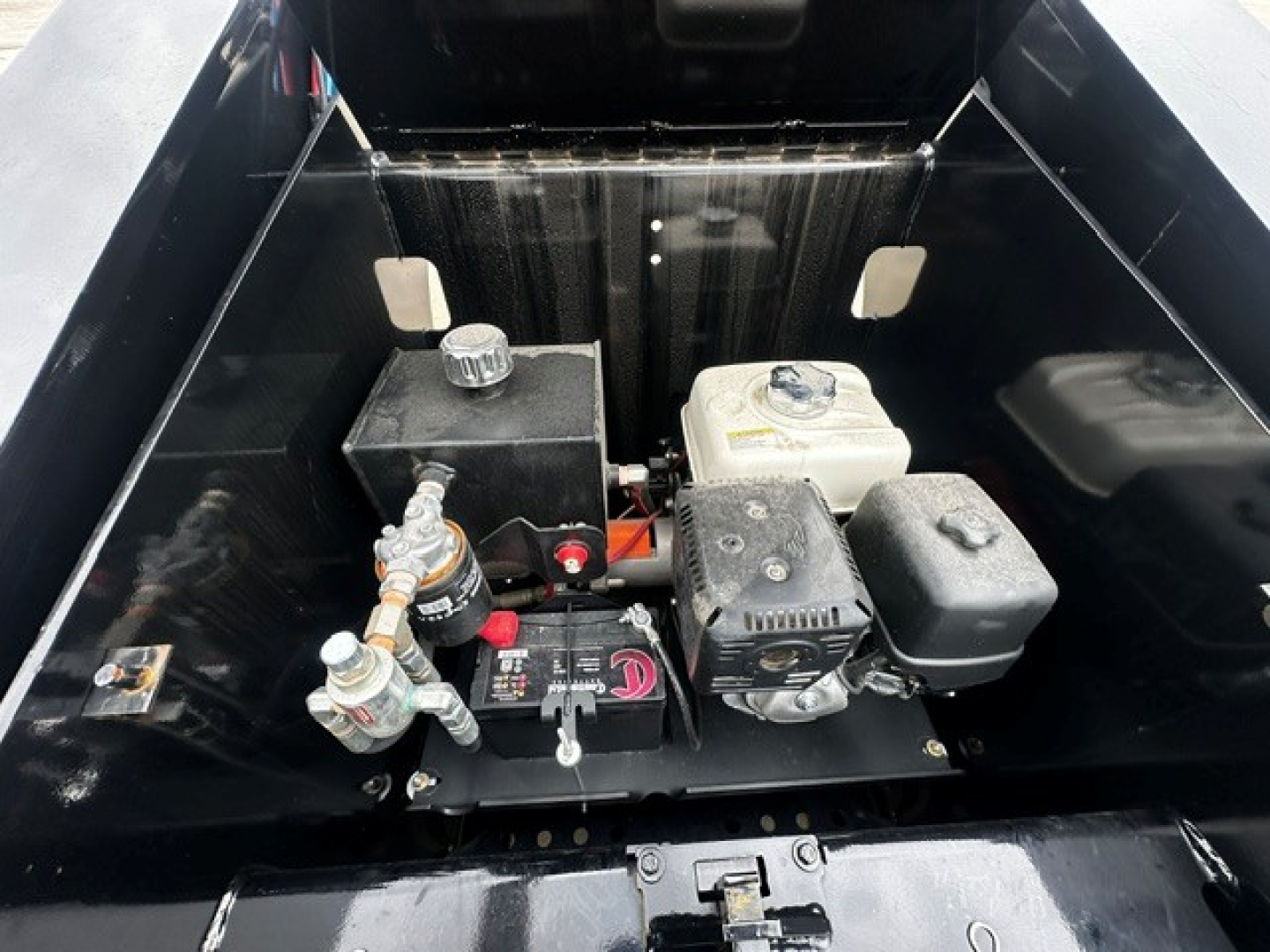 Trailer engine bay showing generator and fluid reservoirs with caps and hoses: black square reservoir, white translucent reservoir, gas engine with pull-start handle, and metal components.