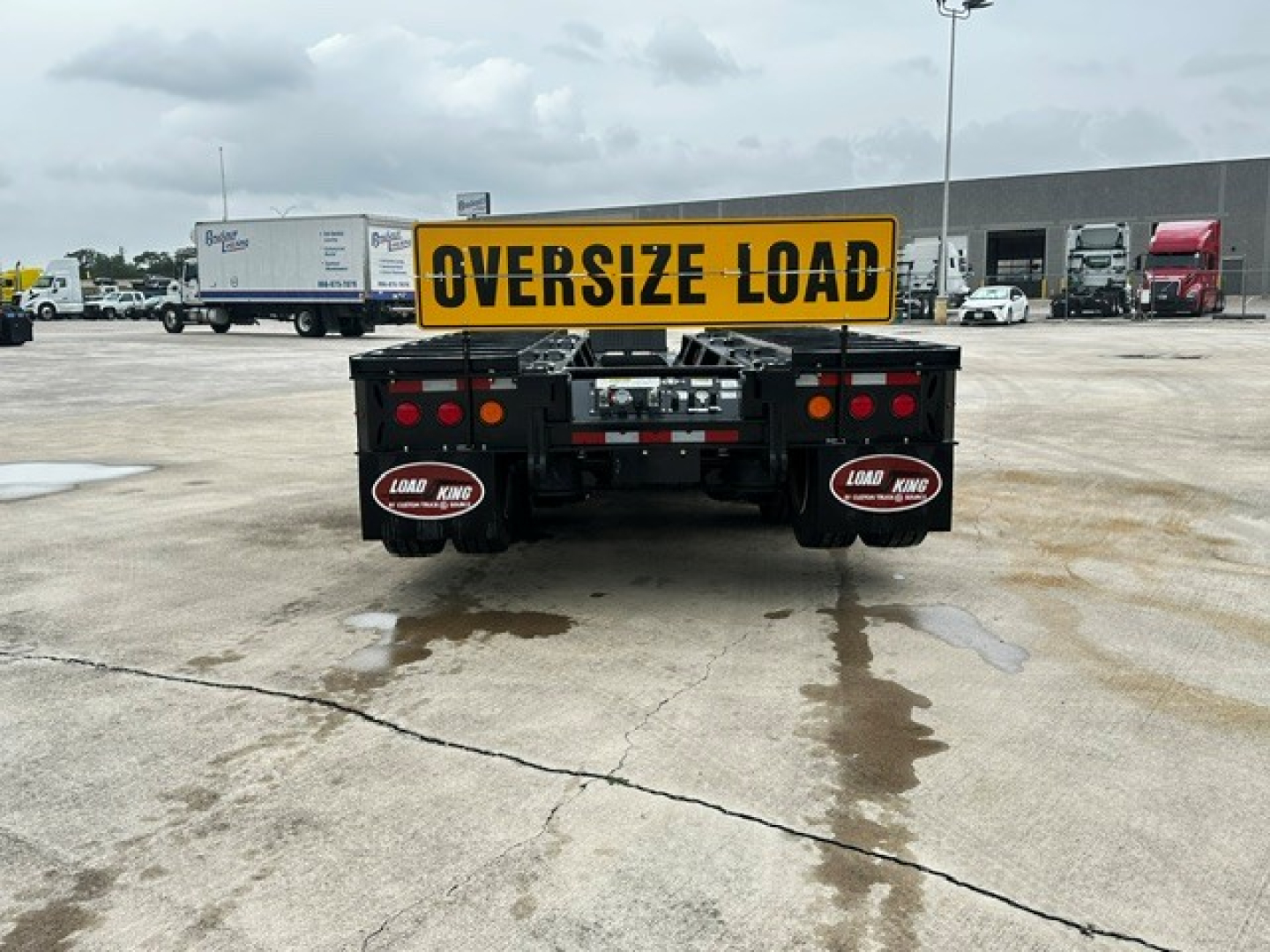 Oversize Load sign mounted on rear of a Load King trailer with taillights, mud flaps, and hydraulic components visible; parking lot with trucks in background.
