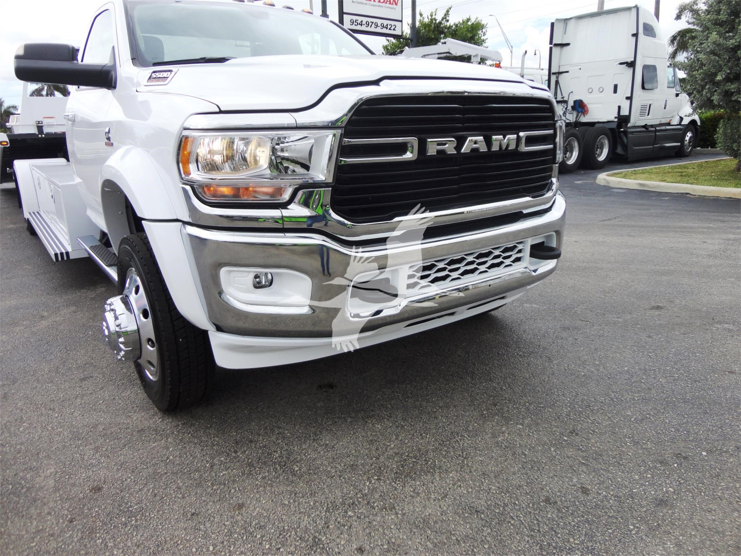 Ram 5500 HD SLT pickup front view with large chrome grille and bumper, white body, rollback truck yard in background.