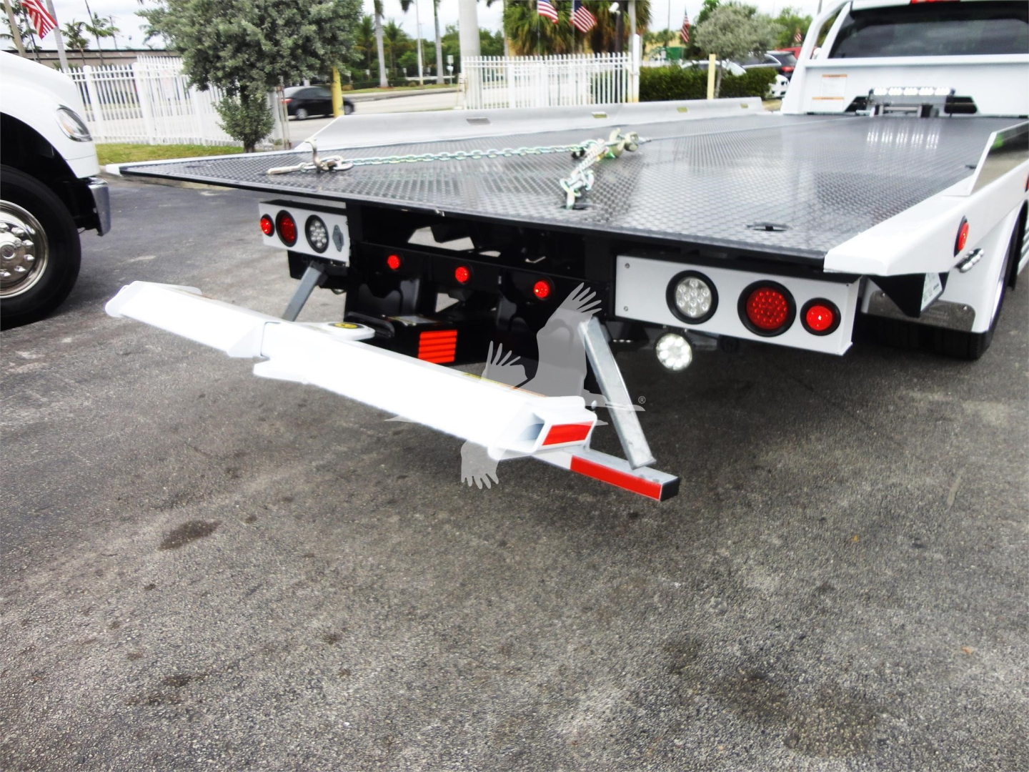Rear view of a white Ram 5500 rollback tow truck with a drop-down lift at the back, diamond plate flatbed, and towing control lights.