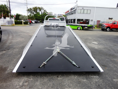 Flatbed tow truck ramp with a dragon logo and tied-down chains in foreground, Ram 5500 chassis visible in distance, dealership lot background.