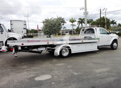 Ram 5500 heavy-duty tow vehicle with flatbed rollback in white, side view showing long bed and wheel chocks.
