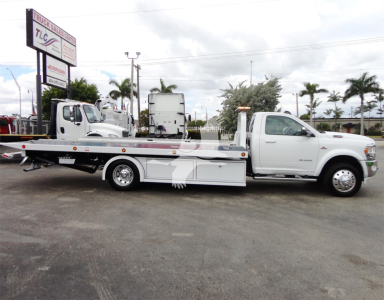 Ram 5500 heavy-duty rollback tow truck side view showing flatbed with hydraulic equipment and white cab, dealership lot in background.