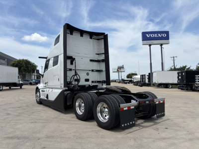 Volvo VNL64T860 tractor rear view showing sleeper cab, dual rear axles, and Volvo mud flaps; visible wheel rims and fuel lines; Volvo sign in background.