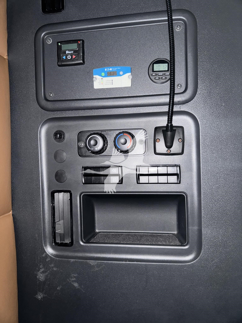 Cab area of a 2023 Freightliner Cascadia 126 dash panel with auxiliary controls and a mounted device; visible are circular knobs, a switch panel, a small digital display, and a storage slot.