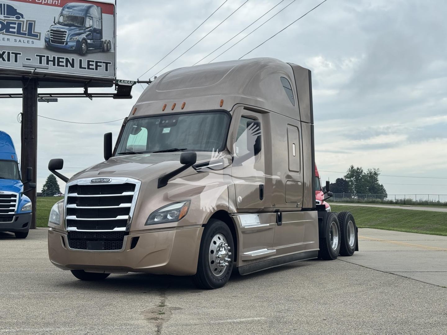 Truck cab in view showing a beige 2023 Freightliner Cascadia 126 sleeper tractor with chrome grille and dual rear axles.