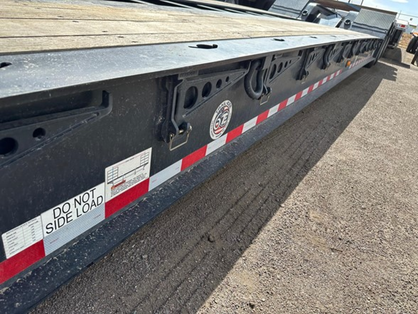 Trailing edge of a lowboy trailer showing Do Not Side Load label, reflective red/white striping, and circular GE logo plate with hooks and fittings along the side.