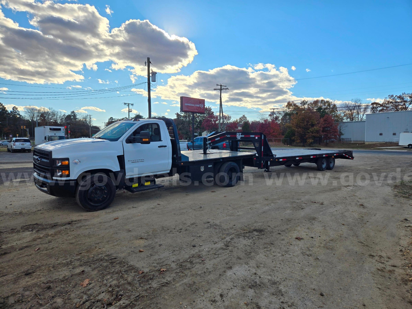 White 2021 Chevrolet medium-duty tow truck with flatbed in a dirt lot; no visible text legible on truck, background shows clouds and trees.
