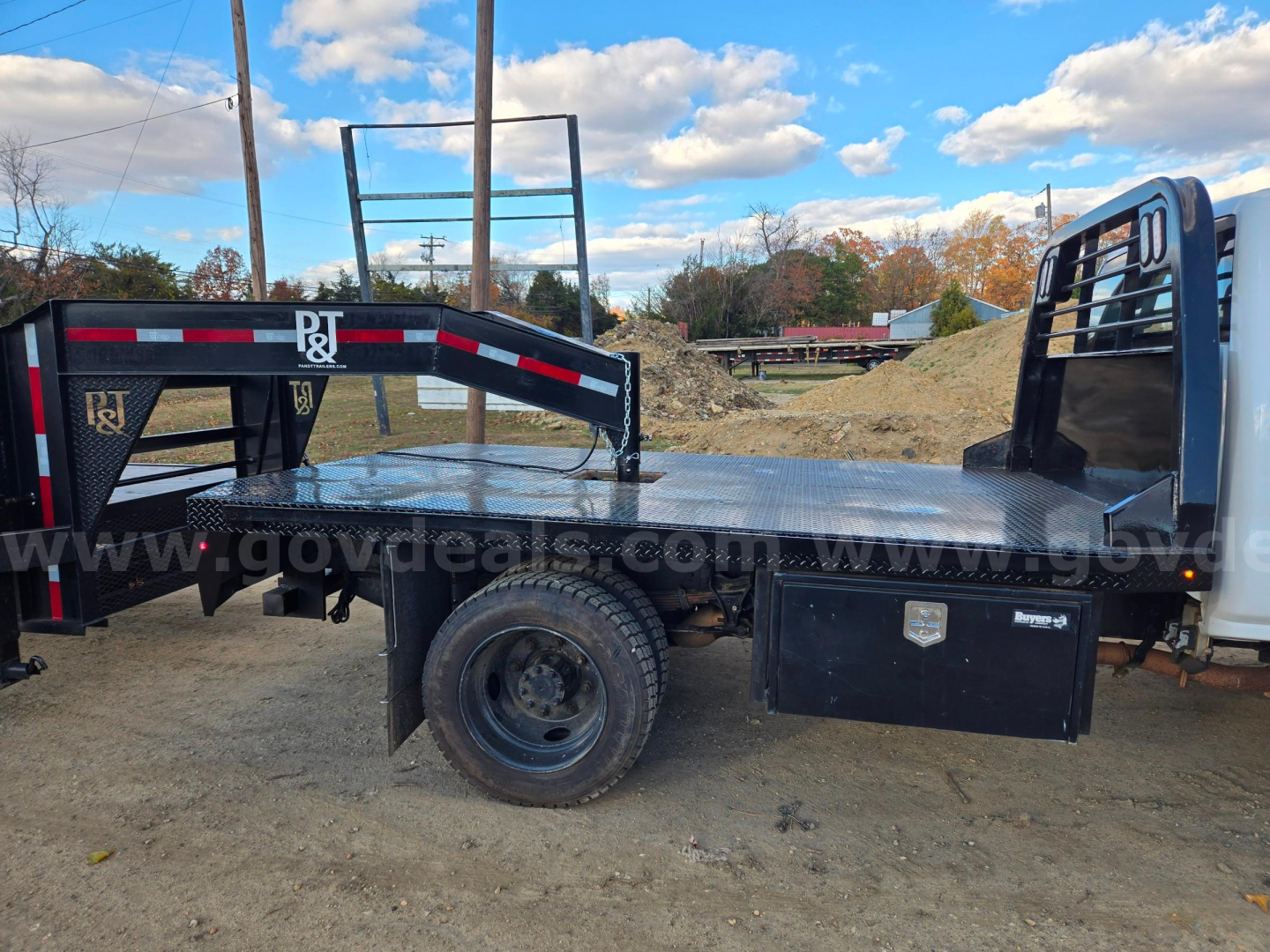 Flatbed truck bed with utility toolbox on passenger side and heavy-duty steel frame, rear flatbed with wheel, side rake rack, and metal ramps in view.