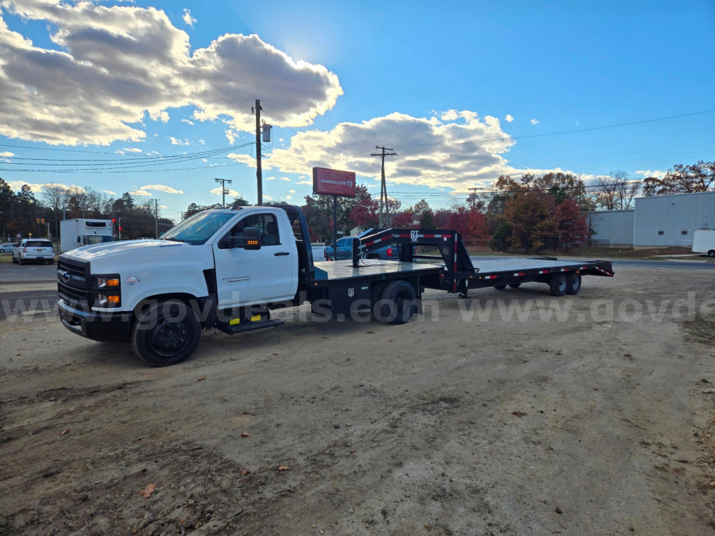 White 2021 Chevrolet medium-duty tow truck with flatbed in a dirt lot; no visible text legible on truck, background shows clouds and trees.