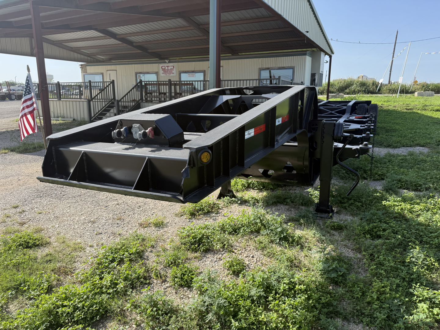 Chassis trailer frame showing black steel box with low profile, several access points and hydraulic connections; American flag and building in background.
