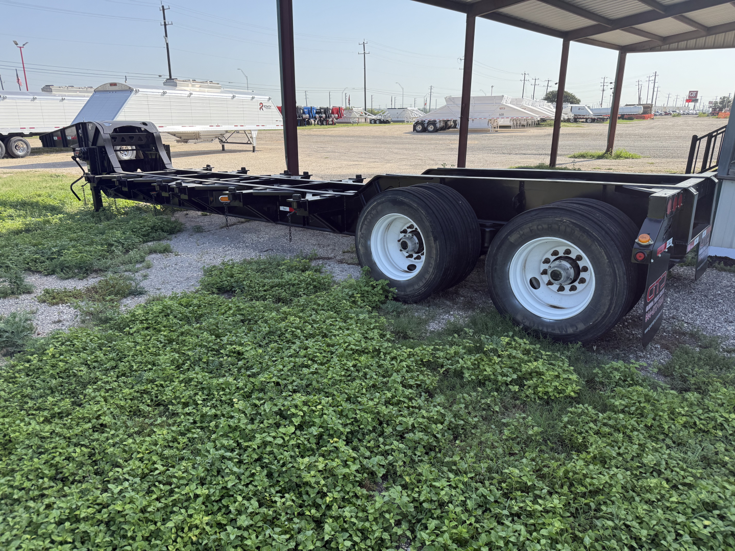 Trailer chassis showing dual rear axles and deck frame under a shelter, with gravel ground and other trailers in background.