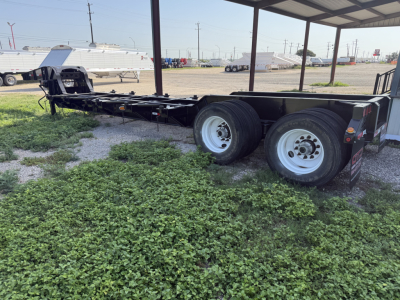 Trailer chassis showing dual rear axles and deck frame under a shelter, with gravel ground and other trailers in background.