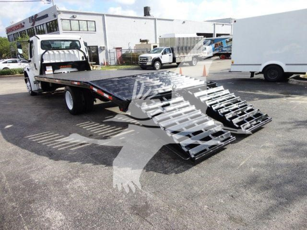 Freightliner M2 106 flatbed truck with a folding aluminum ramp system in the bed, showing a white cab and black flatbed.