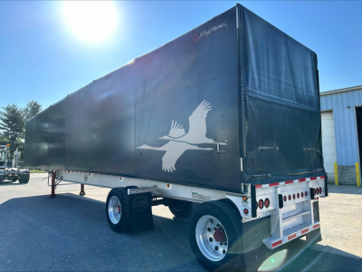 Trailer side view showing 2013 Mac 72K Beam curtainside trailer with dark tarp, white frame, and rear lights; logo of gulls on side, VIN tag not visible.