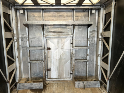 Trailer interior showing a centered metal rear door with surrounding frame and wooden steps on the floor, metal walls with cross members, and a small hatch in the middle door.