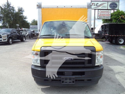 Front view of a yellow 2022 Ford E350 XL SD cargo van with black grille, license plate holder, and large cargo box; dealership signs visible in background with partial text.