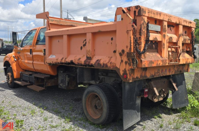 Footage of an orange Ford F-650 dump bed truck with wear on the bed sides, showing the rear and left side. The dump bed has rust-like stains, and a black mudflap is visible behind the rear wheels.