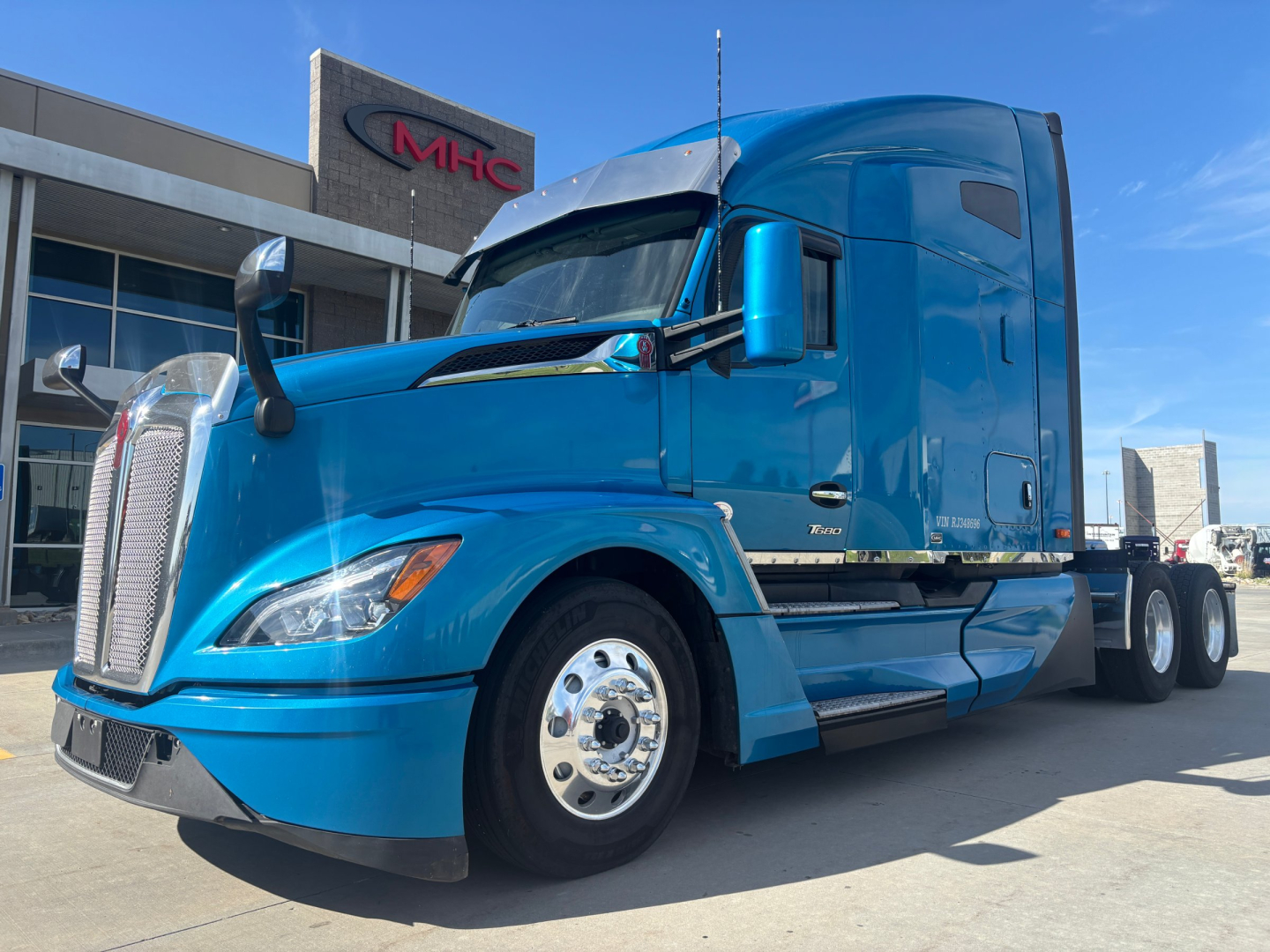 Blue 2024 Kenworth T680 tractor parked outdoors showing front and side view with sleeper cab and chrome wheels.