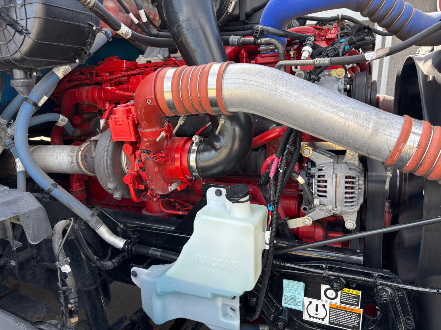 Kenworth T680 engine bay with red engine block, silver turbo hose, blue hoses, and coolant reservoir visible.