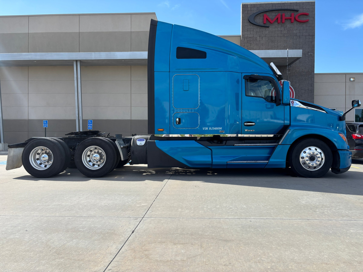 Blue Kenworth T680 sleeper tractor side view showing 6x4 rear axle configuration and MHC dealership building in background.