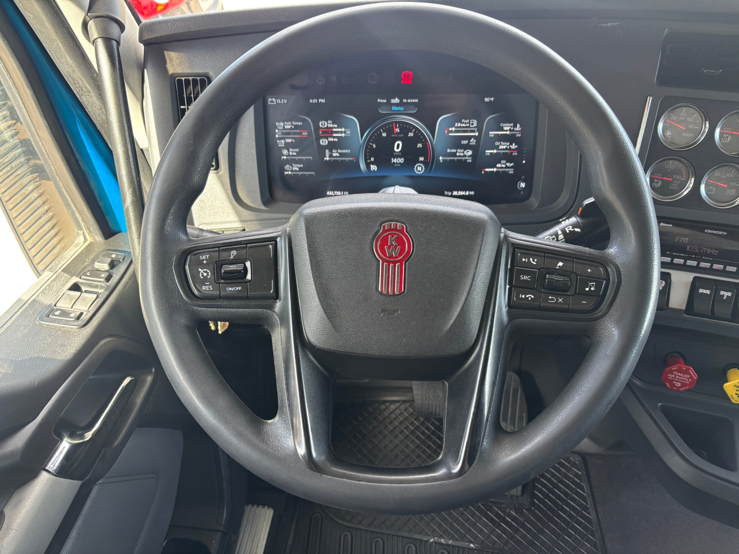 Kenworth truck steering wheel view with digital instrument cluster showing gauges. Steering wheel badge with Kenworth emblem and control buttons on both sides. Dashboard gauges and warning lights visible.