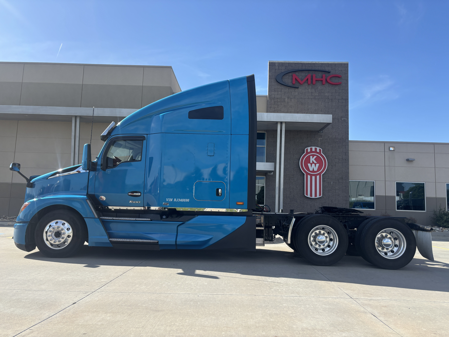 Kenworth T680 semi-truck in blue with sleeper cab, side view showing attached flatbed/construction trailer, company lot in background.