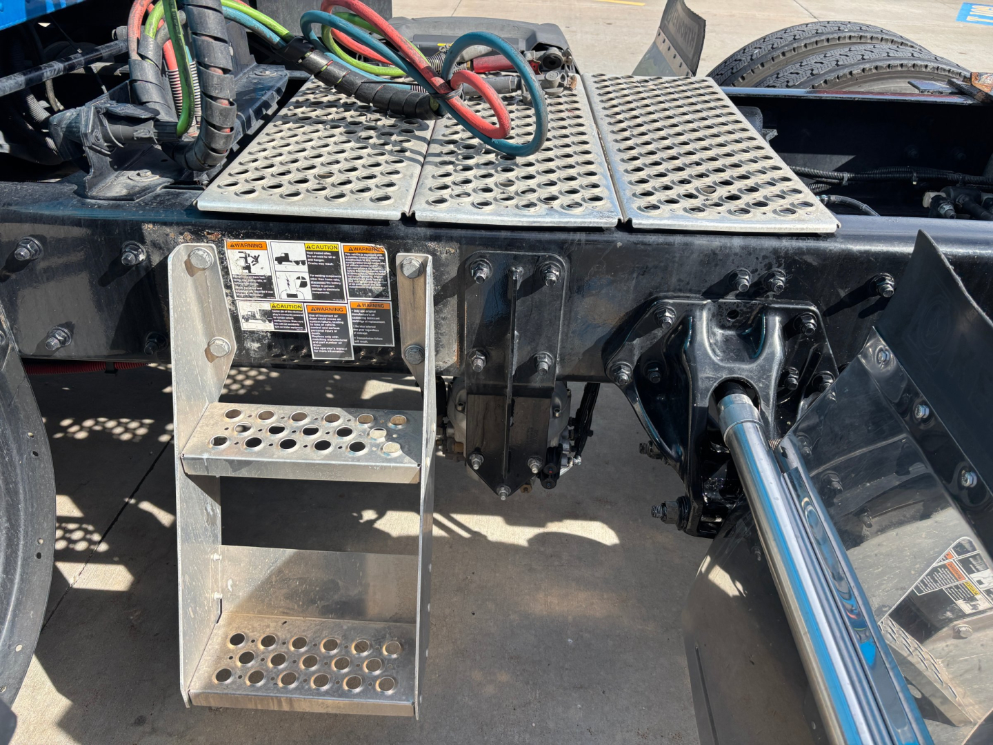 Kenworth T680 undercarriage showing metal steps, hydraulic lines, and a shiny chrome drive shaft; perforated metal platform and control levers visible.