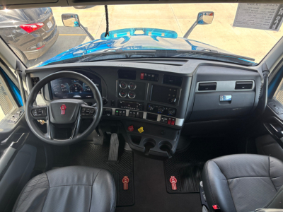 Vehicle cab interior of a 2024 Kenworth T680 showing steering wheel with red logo, dashboard gauges, central controls, and floor mats with red accents.