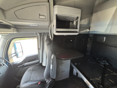 Kenworth T680 cab interior showing overhead sleeper cabinet, microwave, and bench-style seating with a desk; dashboard window and curtains visible