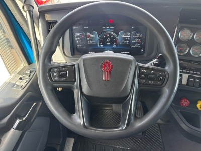 Kenworth truck steering wheel view with digital instrument cluster showing gauges. Steering wheel badge with Kenworth emblem and control buttons on both sides. Dashboard gauges and warning lights visible.