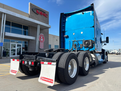 Kenworth T680 tractor with blue sleeper cab, rear view showing third axle, wheels, and mud flaps in a dealership lot.