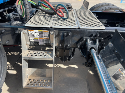 Kenworth T680 undercarriage showing metal steps, hydraulic lines, and a shiny chrome drive shaft; perforated metal platform and control levers visible.