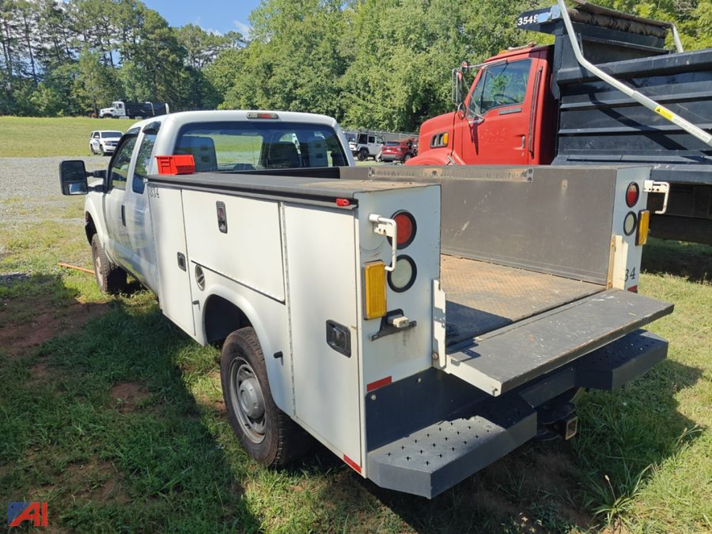 White Ford F-250 utility/work truck with rear deck and toolbox compartments; flatbed with folded tailgate step, dual rear lights, and utility body doors; grassy lot with other trucks in background.