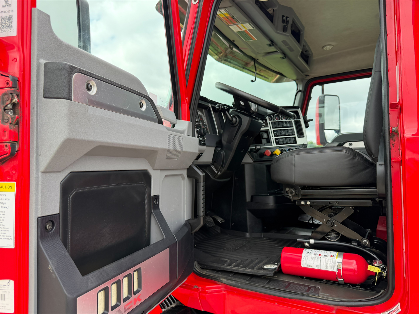 Cab interior of a red Mack AN64T truck showing driver seat, dashboard, steering wheel, and a mounted fire extinguisher under the dash.