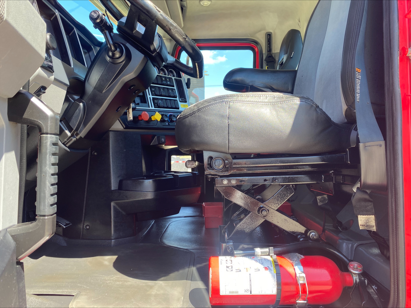 Truck interior showing driver's seat and steering column of a 2021 Mack ANTHEM with a red fire extinguisher mounted near the floor.