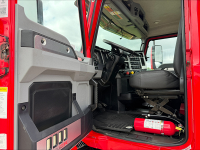 Cab interior of a red Mack AN64T truck showing driver seat, dashboard, steering wheel, and a mounted fire extinguisher under the dash.