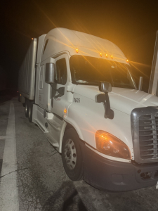 Freightliner Cascadia front-left view showing daytime running lights and front bumper, white exterior, freight trailer connection on sleeper cab, side mirrors.