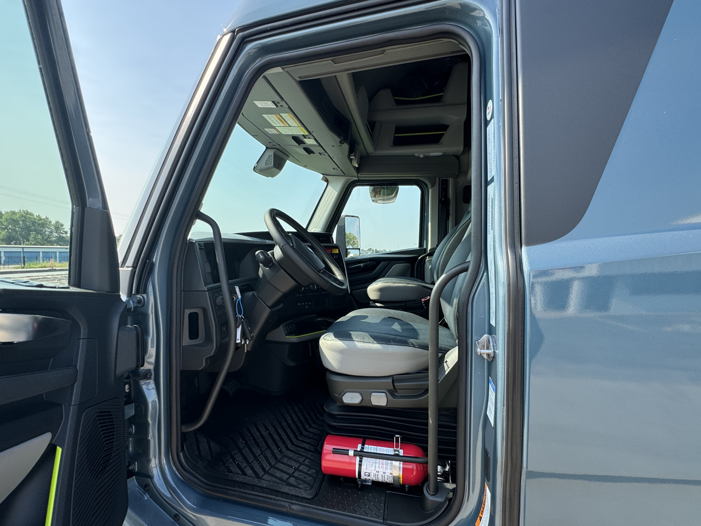 Front cab entry of a 2026 Volvo VNL64T860 truck showing the driver seat and dashboard; Fire extinguisher mounted on the floor near the door.