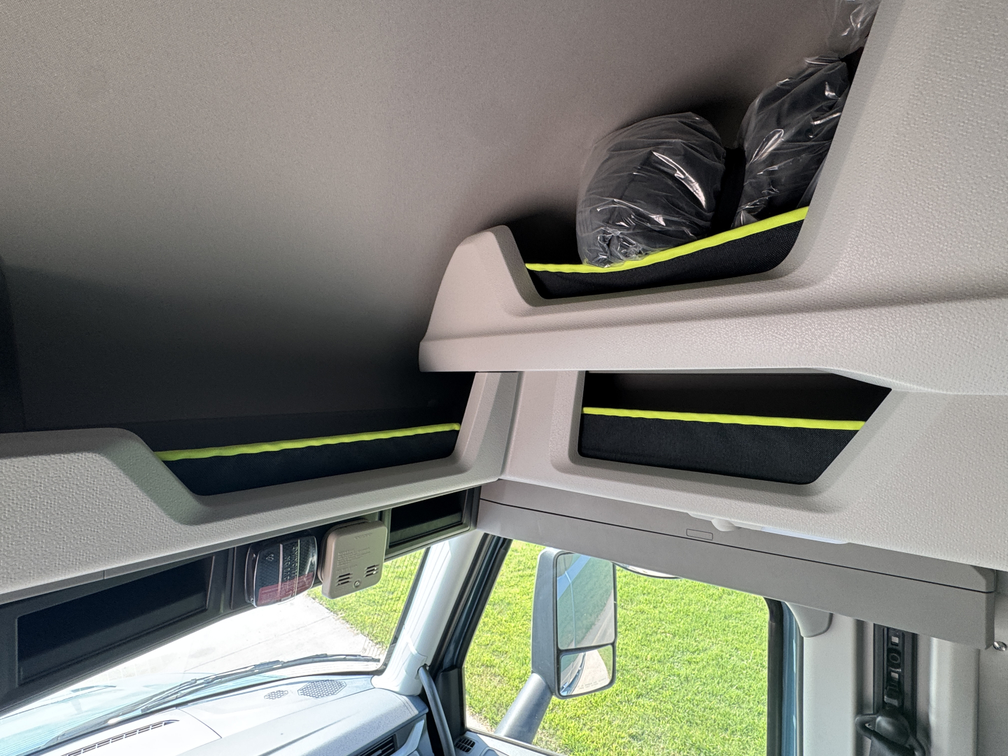 Interior view of a Volvo truck cab showing overhead storage compartments with a seat or bag in black. Light gray interior panels with black trim and a glimpse of the windshield.