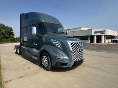 Volvo VNL64T860 sleeper cab truck in blue-gray, front-left view, chrome grille, daytime running lights, tractor-trailer with dual rear axles, dealership building in background.
