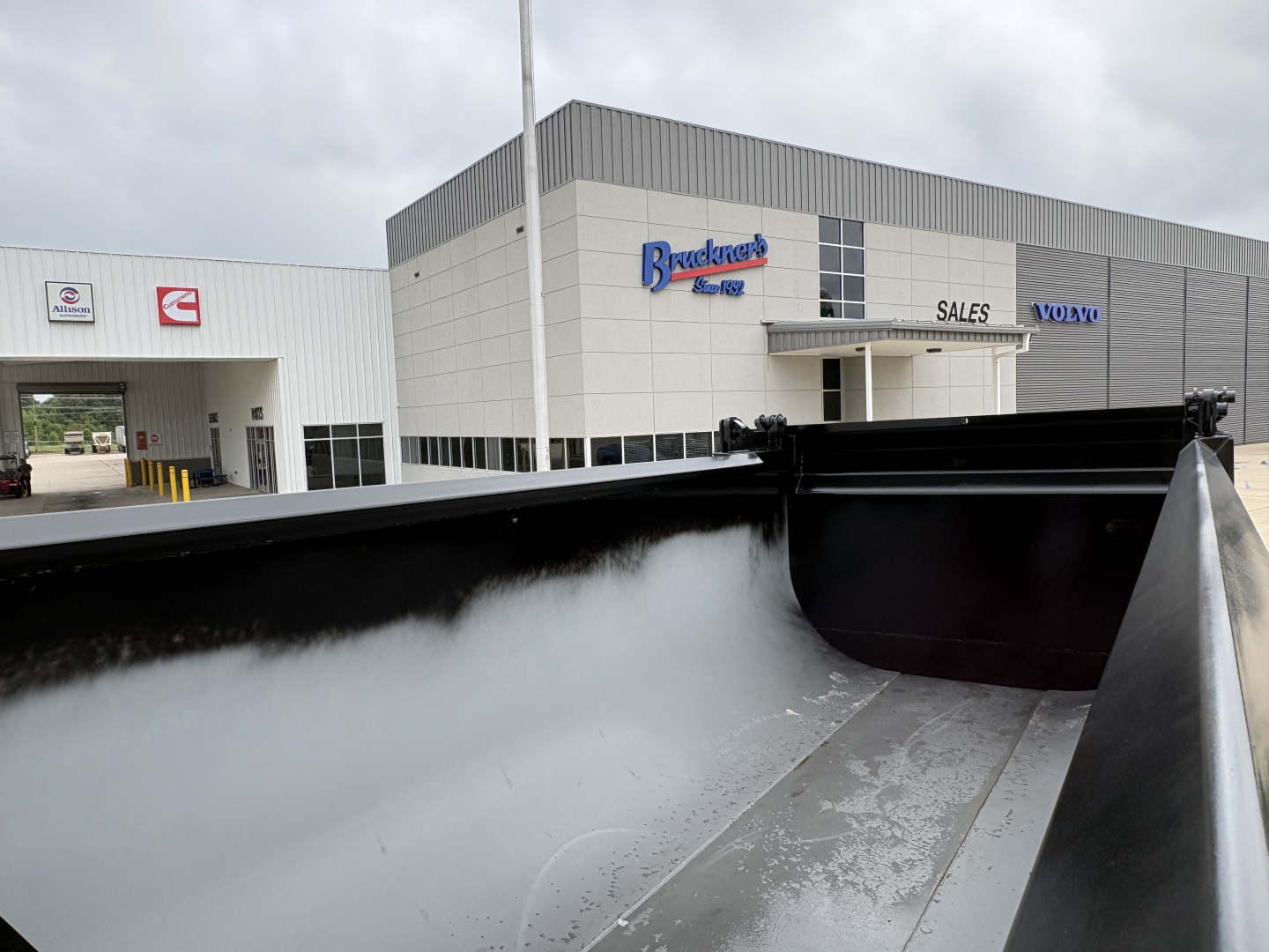 Dump truck bed with high steel sides in foreground and dealership building in background.