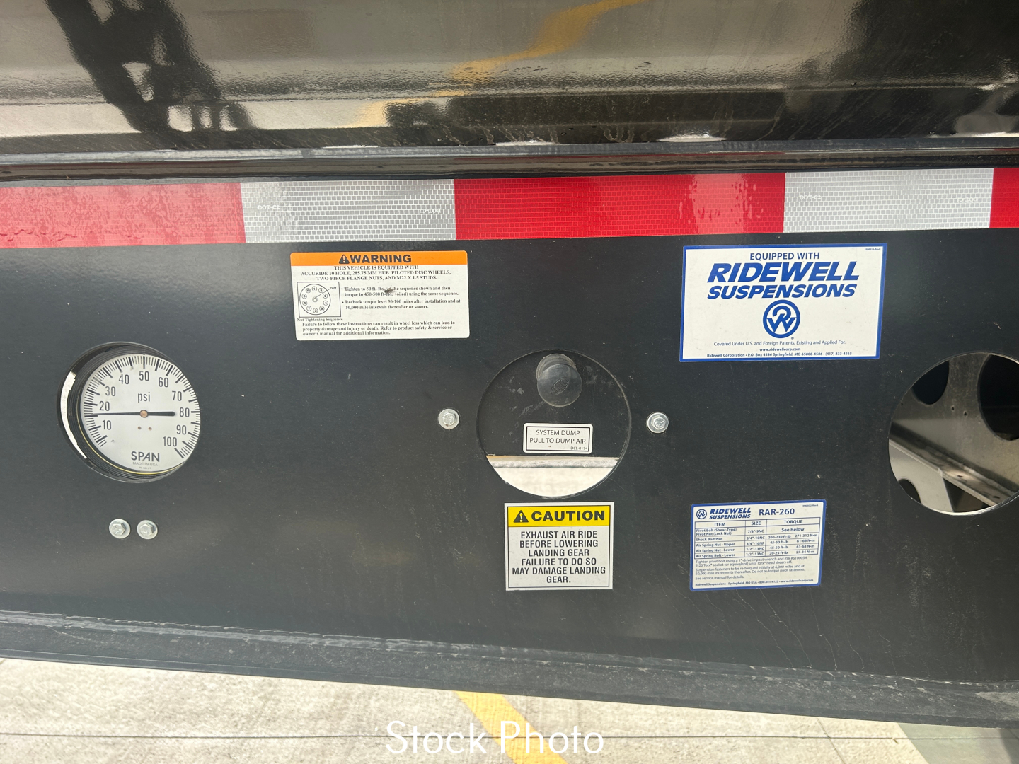 Trailer lower rear panel showing gauges and warning labels. Left gauge reads 0-100 with markings, center circular port with label, several small warning stickers including ‘RIDEWELL SUSPENSIONS’, and a caution label. Stock photo waterma